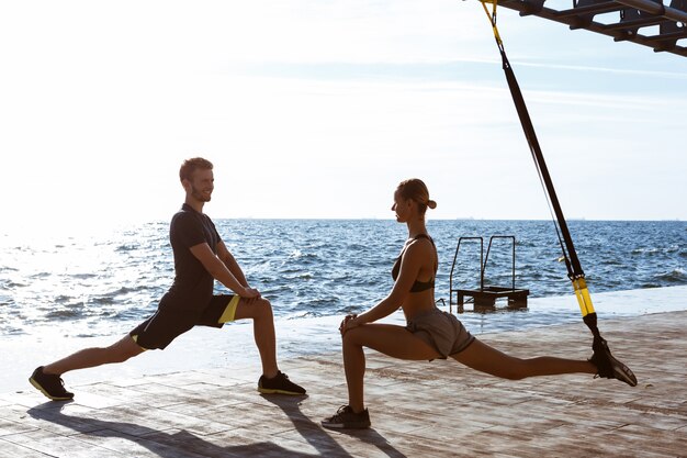 Jóvenes deportivos entrenando con trx cerca del mar en la mañana.