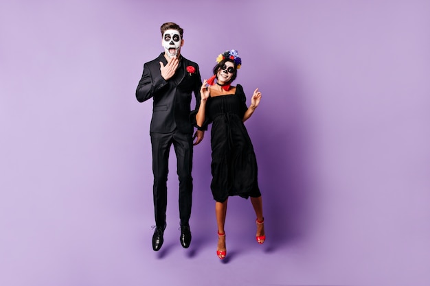 Jóvenes divertidos con arte facial en Halloween posan emocionalmente, saltando sobre fondo púrpura.