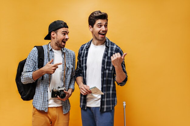 Jóvenes emocionados con camisas a cuadros señalan a la derecha y parecen sorprendidos. Los viajeros tienen boletos y cámara retro en la pared naranja.