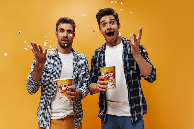 Jóvenes encantadores hombres emocionales esparcen palomitas de maíz sobre fondo naranja Amigos locos en camisas a cuadros posan en aislados