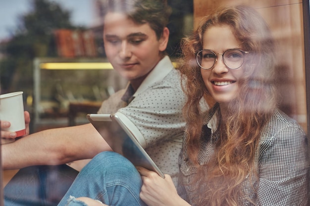 Foto gratuita jóvenes estudiantes felices bebiendo café y usando una tableta digital sentados en un umbral de ventana en un campus universitario durante un descanso.