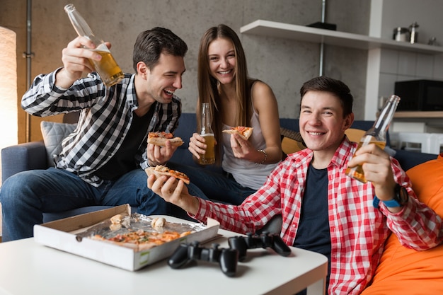 Jóvenes felices comiendo pizza, bebiendo cerveza, divirtiéndose, fiesta de amigos en casa, compañía hipster juntos, dos hombres una mujer, sonriendo, positivo, relajado, pasar el rato, reír,