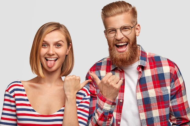 Los jóvenes felices se divierten juntos, se señalan unos a otros con expresiones alegres. Mujer linda divertida muestra la lengua, inconformista masculino barbudo lleno de alegría en camisa a cuadros, aislado sobre la pared blanca