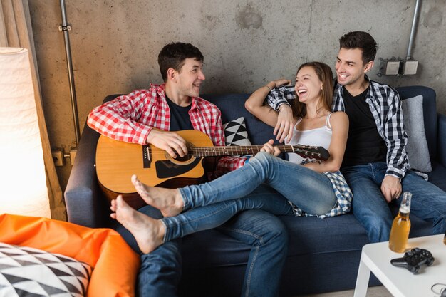 Jóvenes felices divirtiéndose, fiesta de amigos en casa, compañía hipster juntos, dos hombres una mujer, tocando la guitarra, sonriendo, positivo, relajado, bebiendo cerveza, jeans, camisas, estilo casual