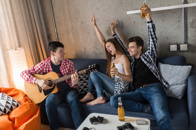 Foto gratuita jóvenes felices divirtiéndose, fiesta de amigos en casa, compañía hipster juntos, dos hombres una mujer, tocando la guitarra, sonriendo, positivo, relajado, bebiendo cerveza