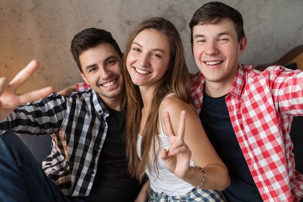 Jóvenes felices haciendo foto divertida selfie, sentados en el sofá, divirtiéndose, fiesta en casa de amigos, compañía hipster juntos, dos hombres una mujer, estilo juvenil, sonriendo, emoción positiva, mirando a puerta cerrada