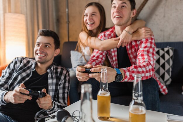 Jóvenes felices jugando videojuegos, divirtiéndose, fiesta de amigos en casa, cerca de las manos sosteniendo el joystick, compañía hipster juntos, sonriendo, positivo, riendo, competencia, botellas de cerveza en la mesa