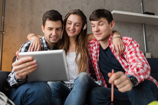 Jóvenes felices mirando en tableta, estudiantes aprendiendo, divirtiéndose, fiesta de amigos en casa, compañía hipster juntos, dos hombres una mujer, sonriendo, positivo, educación en línea, usando el dispositivo