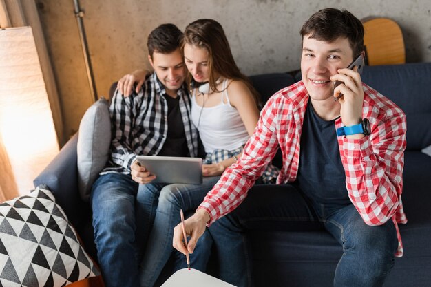 Jóvenes felices usando tableta, estudiantes aprendiendo, divirtiéndose, fiesta de amigos en casa, compañía hipster juntos, dos hombres y una mujer, sonriendo, positivo, educación en línea, hombre hablando por teléfono