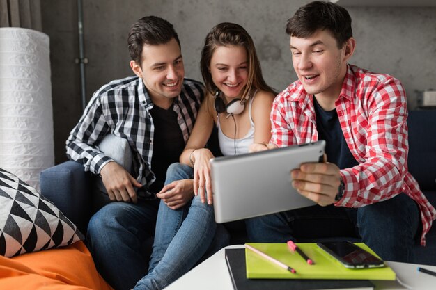 Jóvenes felices usando tableta, estudiantes aprendiendo, divirtiéndose, fiesta de amigos en casa, compañía hipster juntos, dos hombres una mujer, sonriendo, positivo, educación en línea