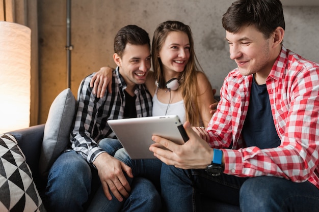 Jóvenes felices usando tableta, estudiantes aprendiendo, divirtiéndose, fiesta de amigos en casa, compañía hipster juntos, dos hombres una mujer, sonriendo, positivo, educación en línea