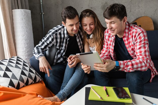 Jóvenes felices usando tableta, estudiantes aprendiendo, divirtiéndose, fiesta de amigos en casa, compañía hipster juntos, dos hombres una mujer, sonriendo, positivo, educación en línea