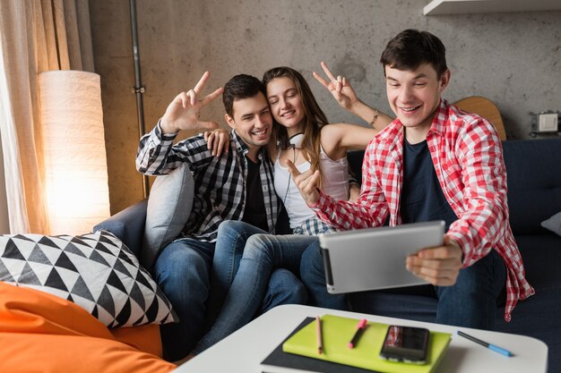 Jóvenes felices usando tableta, estudiantes divirtiéndose, haciendo fotos selfie, fiesta de amigos en casa, compañía hipster juntos, dos hombres y una mujer, sonriendo, positivo, chat en línea