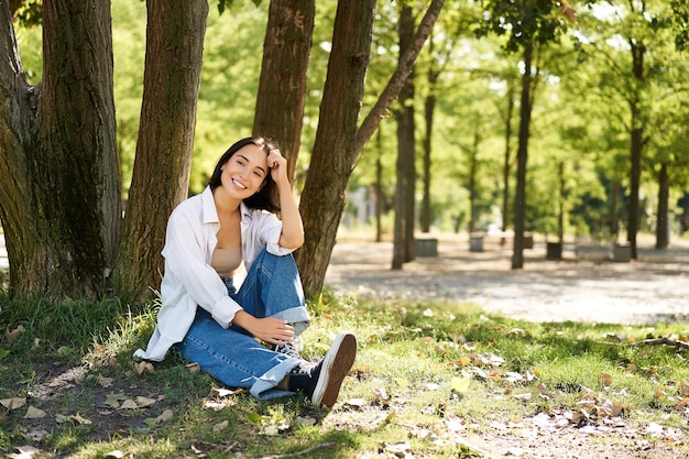 Jóvenes, hermosa chica asiática se sienta cerca de un árbol en el parque y descansa sonriendo y mirando a la distancia