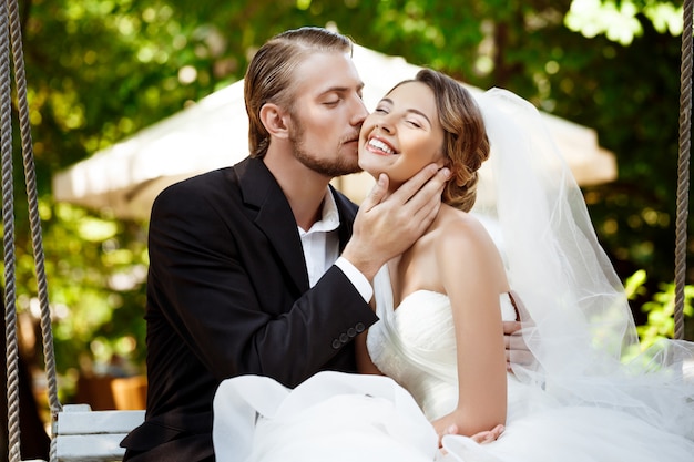 Jóvenes hermosos recién casados sonriendo, besando, sentado en el columpio en el parque.