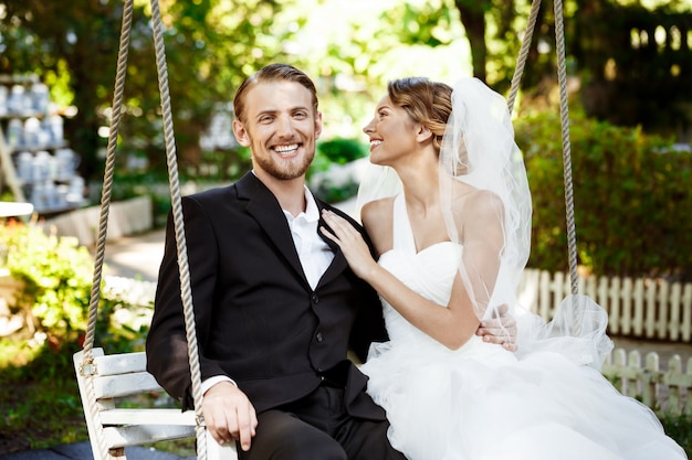Jóvenes hermosos recién casados sonriendo, riendo, sentado en el columpio en el parque.