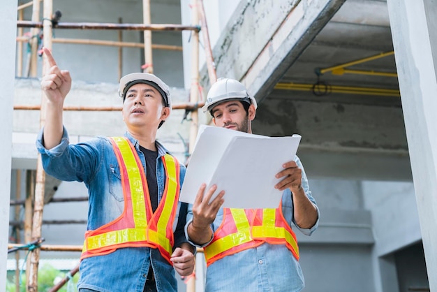 Jóvenes ingenieros asiáticos y colegas ingenieros caucásicos en el inspector del sitio y discuten juntos usando una computadora portátil y un plano de papel en el concepto de ideas de renovación del sitio de construcción del edificio