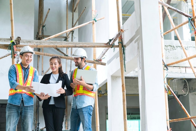 Los jóvenes ingenieros asiáticos trabajan en equipo como inspectores del sitio y discuten juntos usando una computadora portátil y un plano de papel en el concepto de ideas de renovación del sitio de construcción del edificio