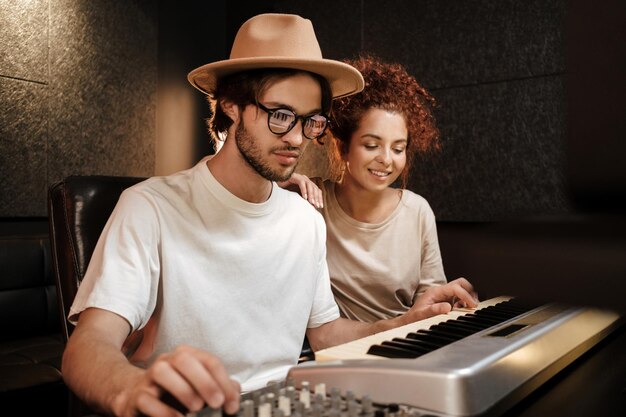 Jóvenes músicos con estilo felizmente grabando una nueva canción en el teclado del piano en un estudio de sonido moderno. Atractivo hombre y mujer creativos trabajando en un nuevo álbum de música
