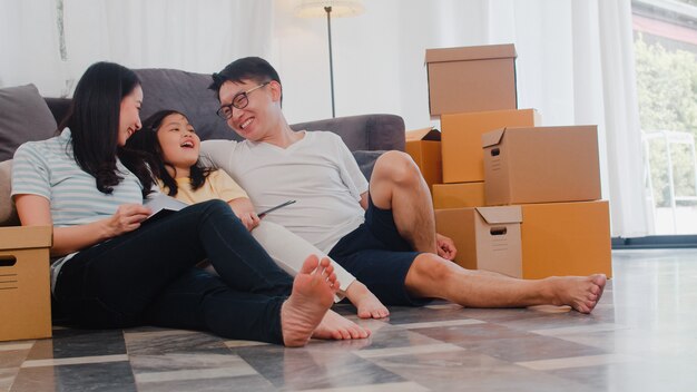 Los jóvenes propietarios de viviendas asiáticos felices compraron una casa nueva. China mamá, papá e hija abrazados mirando hacia el futuro en un nuevo hogar después de mudarse en reubicación sentados en el piso con cajas juntas.