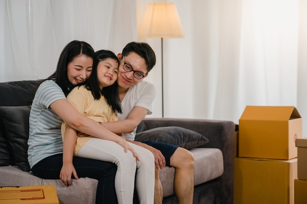 Los jóvenes propietarios de viviendas asiáticos felices compraron una casa nueva. Los japoneses mamá, papá e hija se abrazan mirando hacia el futuro en un nuevo hogar después de mudarse en reubicación sentados juntos en un sofá con cajas.