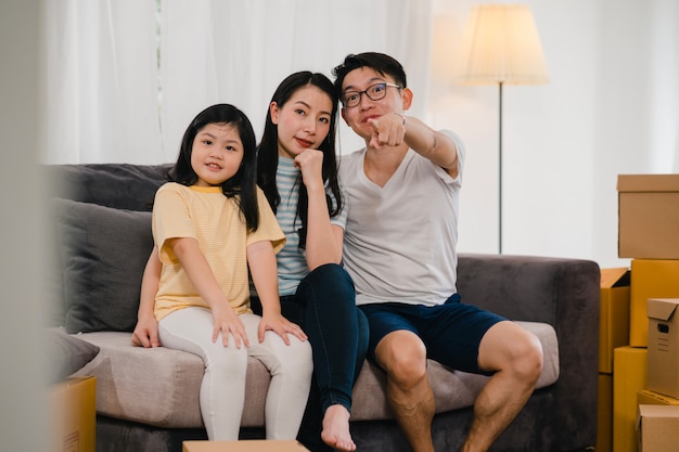 Los jóvenes propietarios de viviendas asiáticos felices compraron una casa nueva. Los japoneses mamá, papá e hija se abrazan mirando hacia el futuro en un nuevo hogar después de mudarse en reubicación sentados juntos en un sofá con cajas.