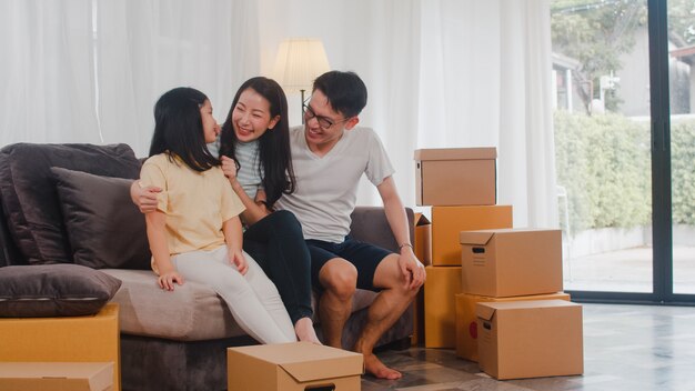 Los jóvenes propietarios de viviendas asiáticos felices compraron una casa nueva. Los japoneses mamá, papá e hija se abrazan mirando hacia el futuro en un nuevo hogar después de mudarse en reubicación sentados juntos en un sofá con cajas.