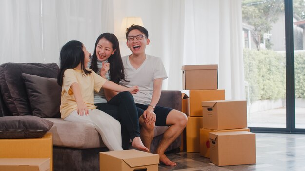 Los jóvenes propietarios de viviendas asiáticos felices compraron una casa nueva. Los japoneses mamá, papá e hija se abrazan mirando hacia el futuro en un nuevo hogar después de mudarse en reubicación sentados juntos en un sofá con cajas.