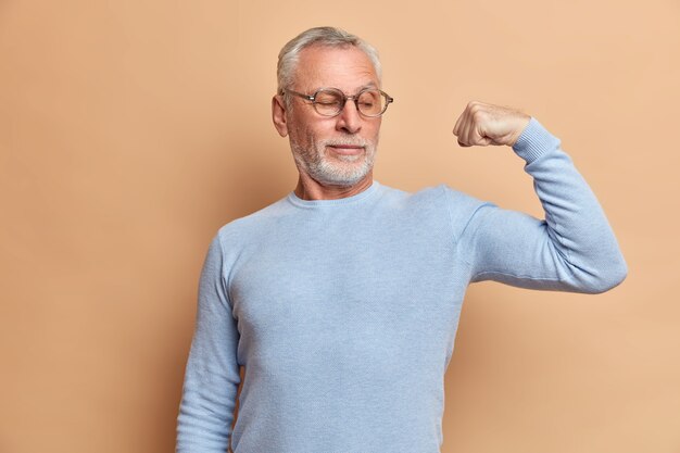 El jubilado hombre de pelo gris barbudo fuerte muestra bíceps y se para con el brazo levantado en el interior usa un jersey y gafas dice mira mi fuerza demuestra músculos aislados sobre una pared marrón