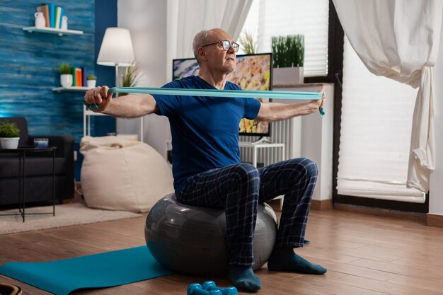 Jubilado senior hombre sentado sobre una pelota suiza en la sala de estar haciendo ejercicios de fitness sanitario estirando los músculos del brazo con banda elástica de resistencia. La fuerza del cuerpo de entrenamiento de pensionista en la sala de estar