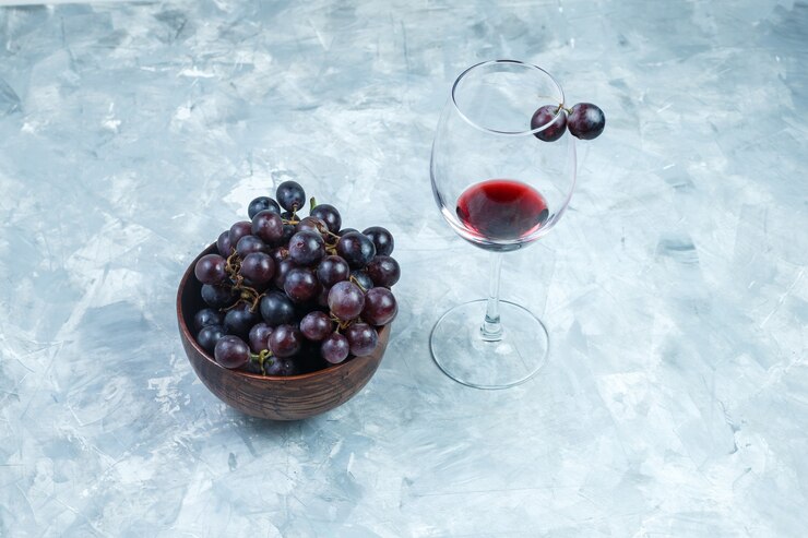 Foto gratuito juego de una copa de vino y uvas negras en un cuenco de barro sobre un fondo gris sucio. vista de ángulo alto.