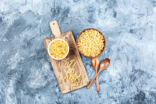 Juego de cucharas de madera y pasta variada en tazones sobre un fondo gris de yeso y tabla de cortar. vista superior.