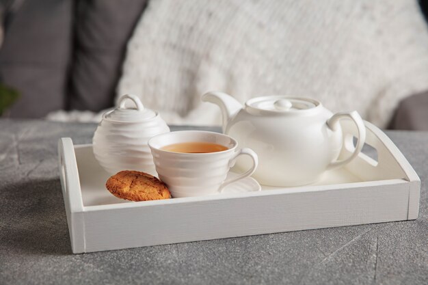 Juego de té blanco y tortas en mesa de madera gris. Bandeja de madera blanca con tazas, tetera y guirnaldas de luces.