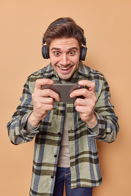 Jugador adicto optimista juega videojuegos en el teléfono inteligente mira la pantalla del celular se alegra de usar auriculares inalámbricos en los oídos vestidos con camisa a cuadros aislada sobre fondo beige