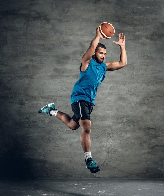 Un jugador de baloncesto negro saltando con ropa deportiva azul sobre fondo gris.