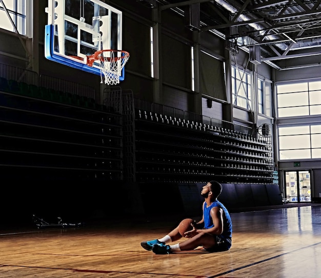 El jugador de baloncesto profesional negro se sienta en un campo bajo el aro en una sala de juegos.