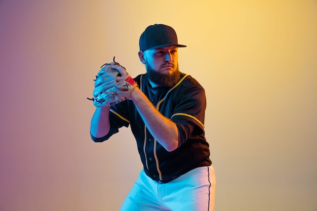 Jugador de béisbol, lanzador con uniforme negro practicando y entrenando en pared degradada con luz de neón. Joven deportista profesional en acción y movimiento. Estilo de vida saludable, deporte, concepto de movimiento.