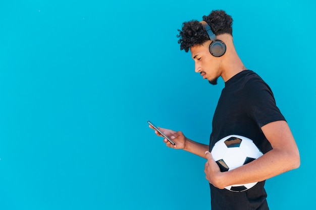 Jugador étnico con fútbol usando teléfono y auriculares.