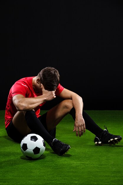 Jugador de fútbol sentado con la pelota en el césped, partido perdido