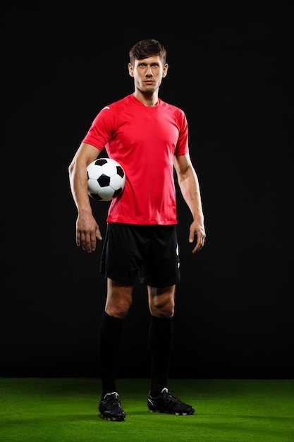 Jugador de fútbol en uniforme negro y rojo con balón en la mano de pie sobre el césped sobre la pared negra, mirando al frente