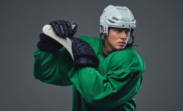 Jugador de hockey con equipo de protección verde y casco blanco de pie con el palo de hockey sobre un fondo gris.