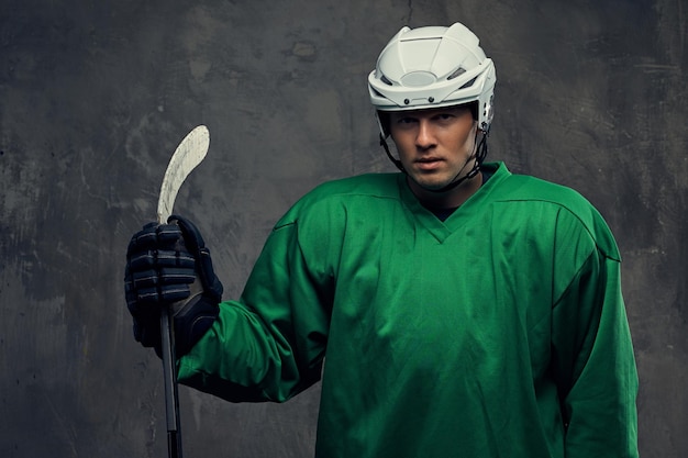 Jugador de hockey con equipo de protección verde y casco blanco de pie con el palo de hockey sobre un fondo gris.