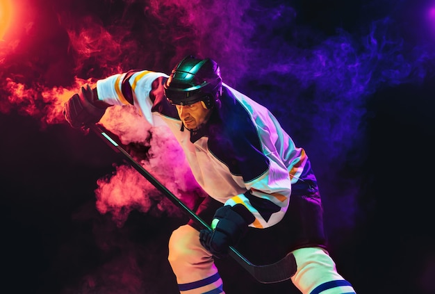 Jugador de hockey masculino con el palo en la cancha de hielo y la pared de color neón oscuro