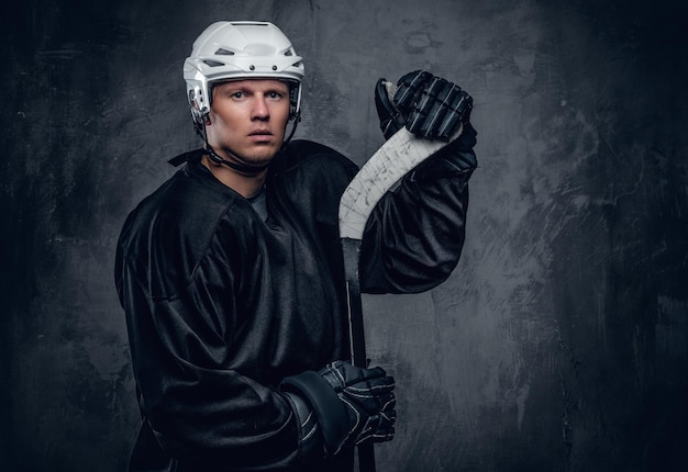 El jugador de hockey con ropa deportiva negra y casco blanco sostiene el palo de juego sobre fondo gris.