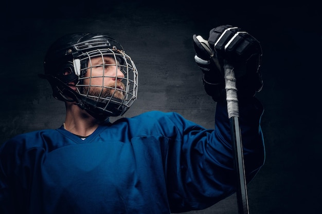 Un jugador de hockey sobre hielo barbudo en una ropa deportiva azul sostiene un palo de juego en iluminación de contraste sobre fondo de viñeta gris.
