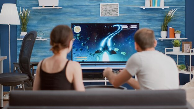 Jugador jugando consola de videojuegos tirador en primera persona en la televisión mientras la novia come palomitas de maíz y le da consejos sentados en el sofá. Pareja en el sofá disfrutando de la simulación de juegos en la sala de estar moderna.