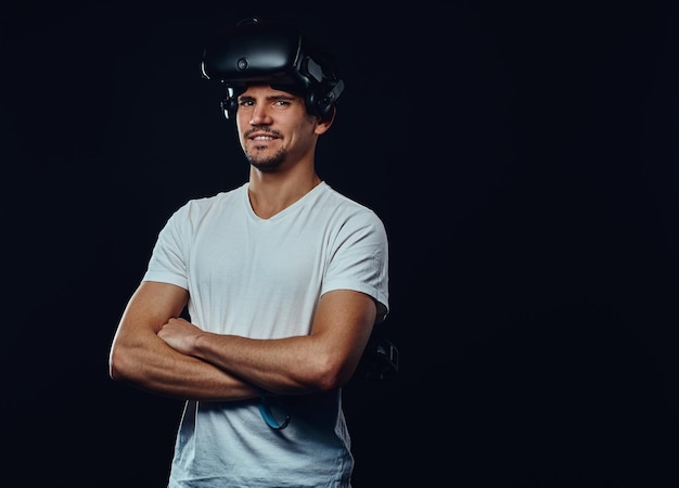 Jugador profesional con cerdas vestido con camisa blanca con gafas de realidad virtual y joysticks, posando con los brazos cruzados. Aislado sobre fondo oscuro.