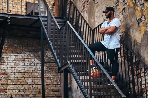 Un jugador de streetball barbudo afroamericano con gorra vestido con ropa deportiva sostiene una pelota de baloncesto en las escaleras sostiene una pierna en una pelota en un gueto.