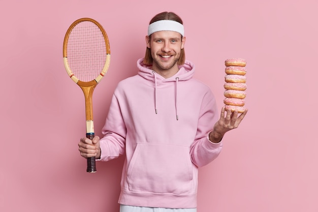 El jugador de tenis feliz elige entre un estilo de vida saludable y la comida dañina sostiene la raqueta y la pila de donas dulces usa una sudadera y una diadema. Hombre barbudo europeo va a jugar al bádminton