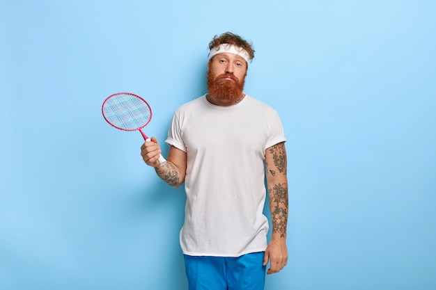 Jugador de tenis pelirrojo cansado sostiene la raqueta mientras posa contra la pared azul